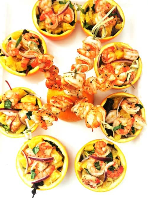 Shrimp salad in orange peel cups on white plate with shrimp skewers