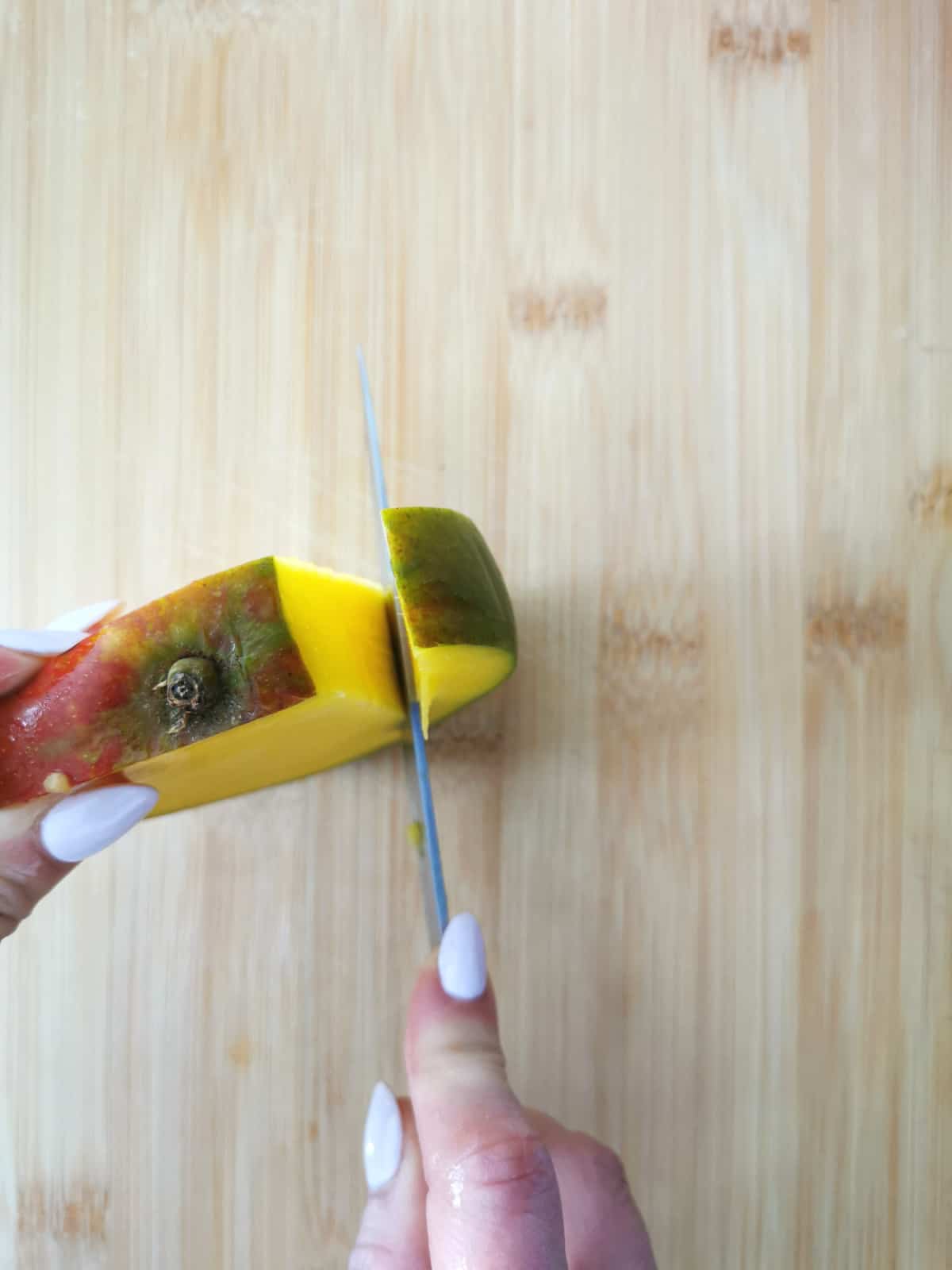 hand holding a knife cutting side of mango to trim flesh from pit.