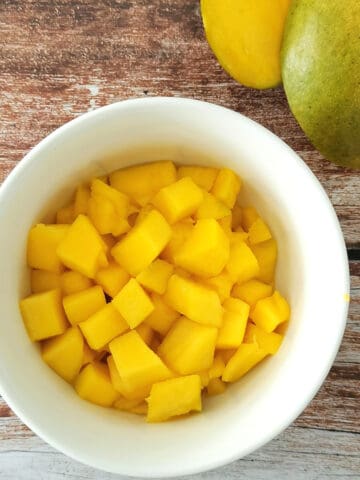 cut up mango in a small white bowl with large mango pieces on the side.