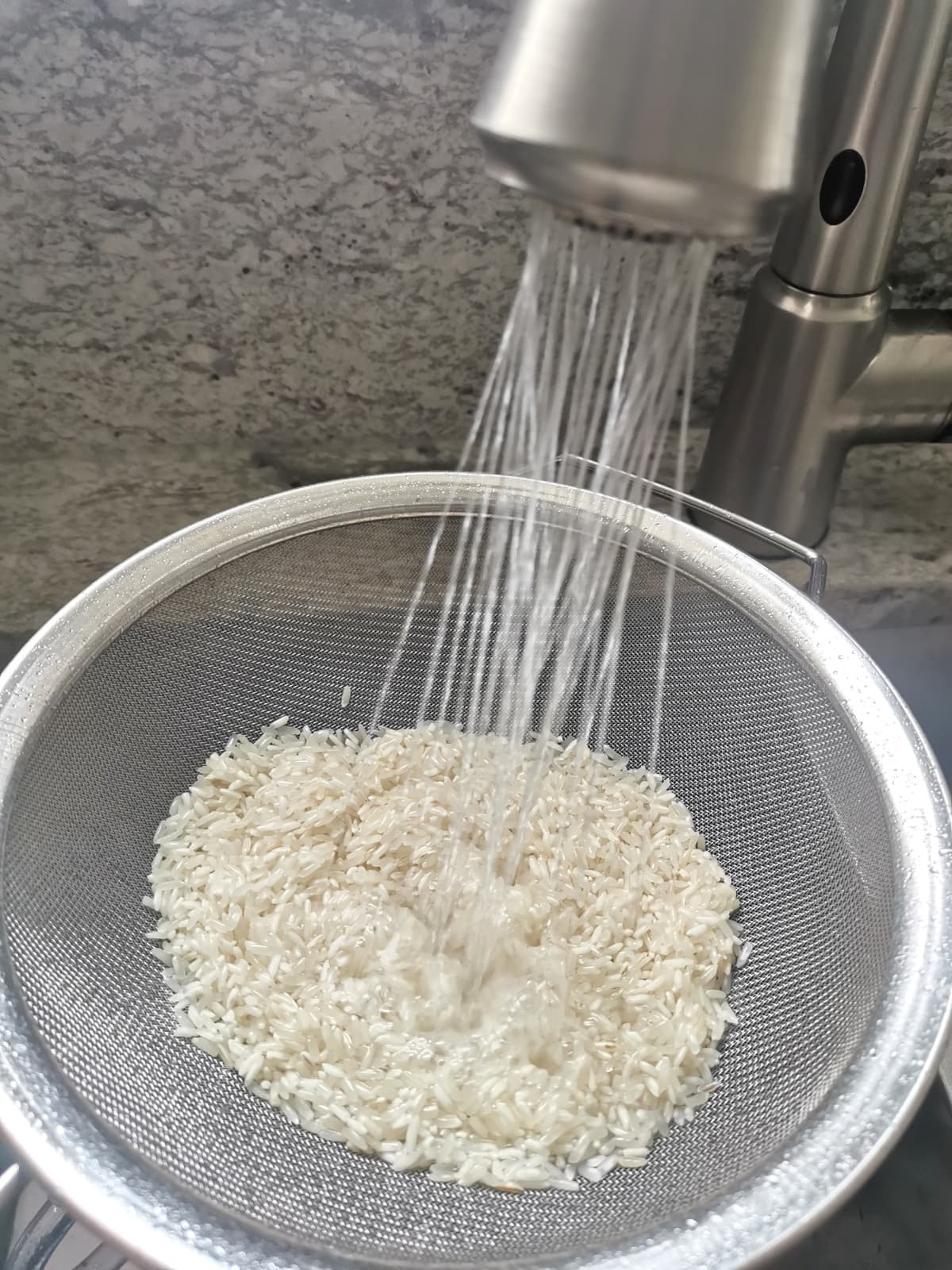 white rice in a mesh strainer rinsing under running water in the sink.