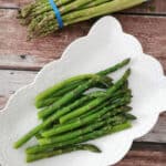 blanched asparagus in a white serving plate with raw asparagus on the side.
