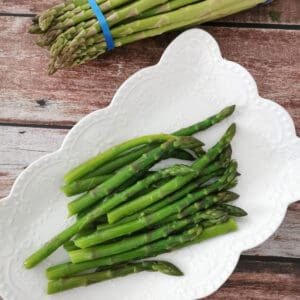 blanched asparagus in a white serving plate with raw asparagus on the side.