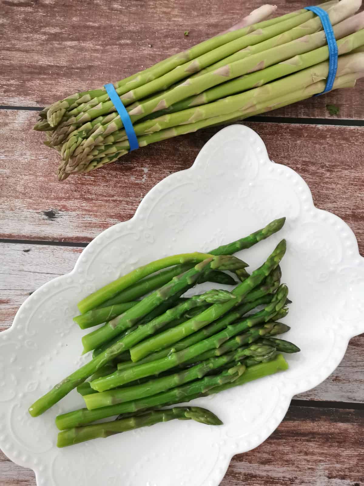 blanched asparagus in a white serving plate with raw asparagus on the side.