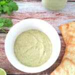 Cilantro lime tahini sauce in a white bowl with crackers, lime and cilantro on the side.