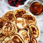 Russian Pancakes Oladi on a large white plate with berries and maple syrup on the side.