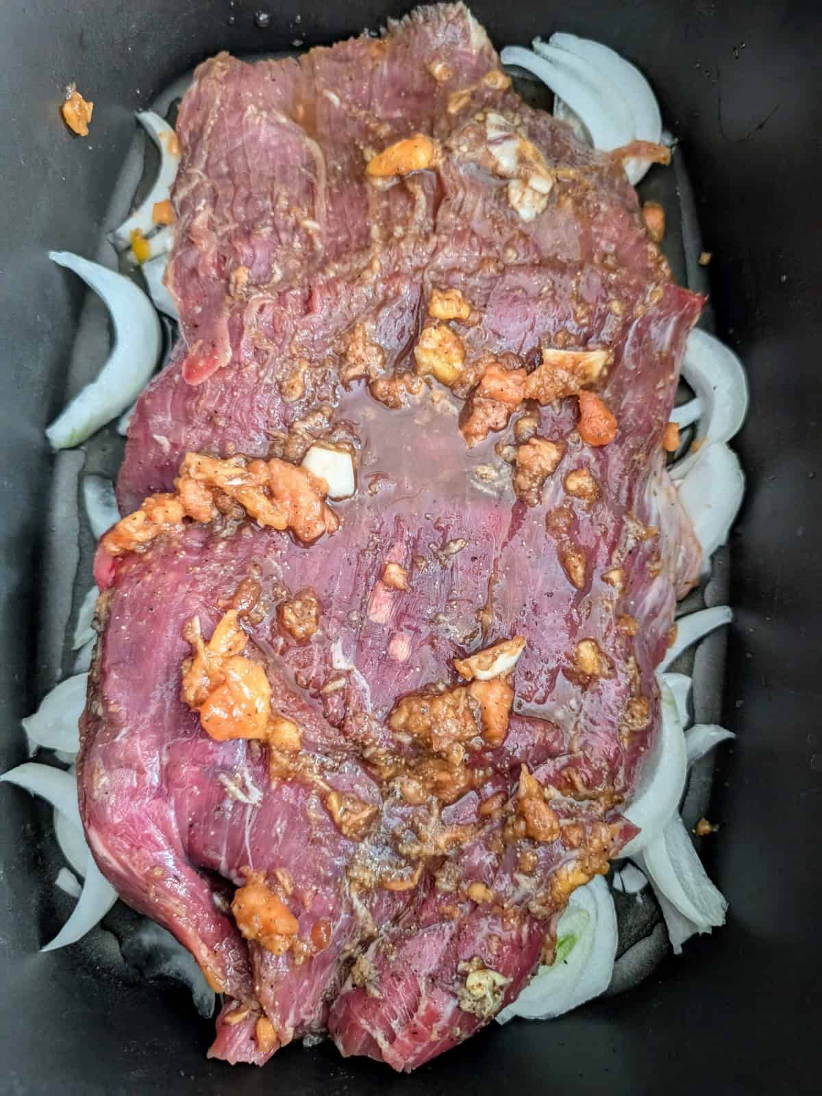 seasoned flank steak placed over sliced onions in a slow cooker.