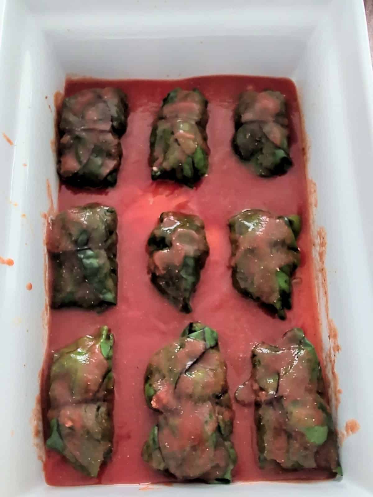 Swiss chard rolls placed in a baking dish and topped with tomato sauce.