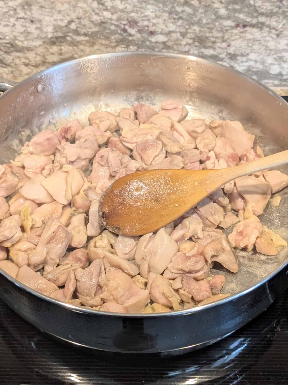 cut-up chicken thighs cooking in a large stainless steel skillet with wooden spoon.