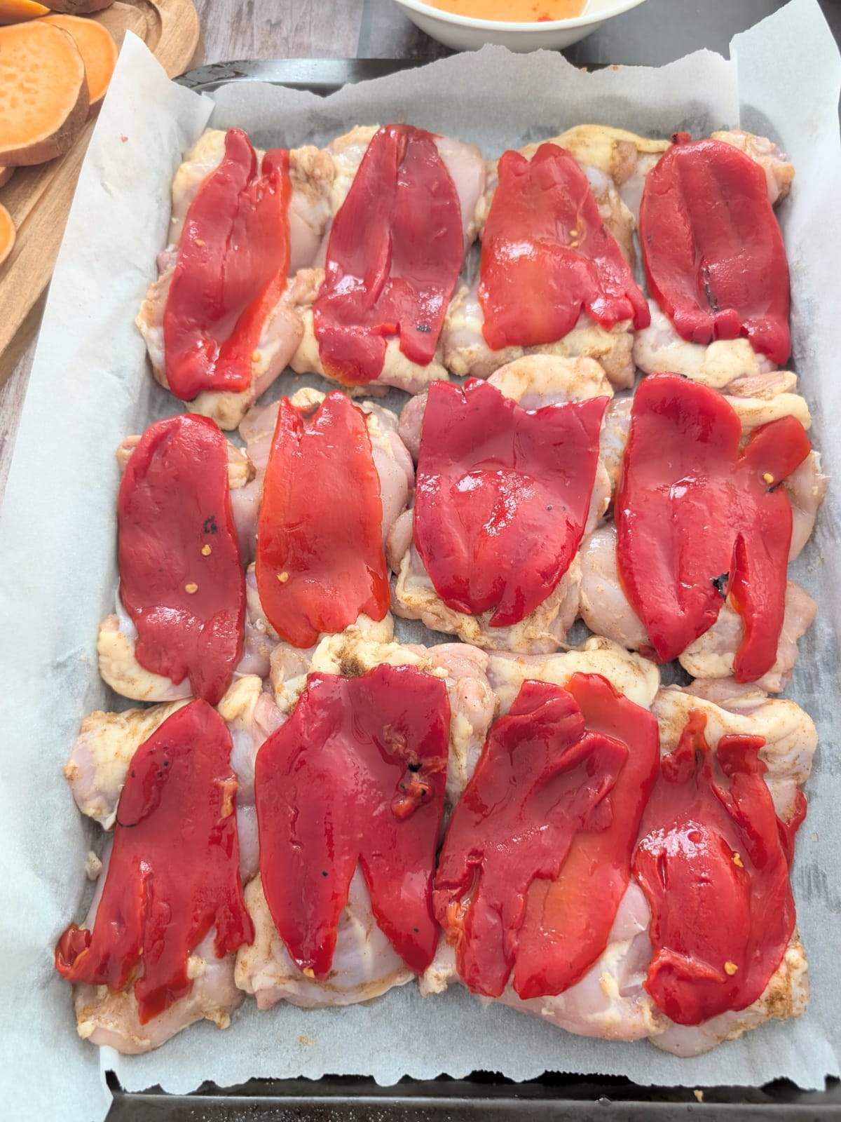 Seasoned chicken thighs topped with jarred roasted red peppers on a baking sheet.