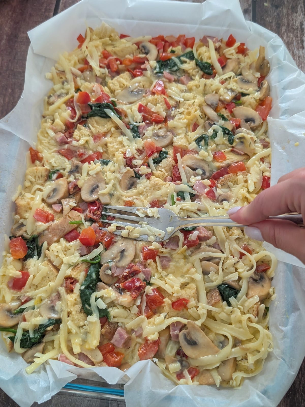 Egg and milk custard poured over bread, vegetables, ham and cheese for breakfast strata and fork pressing down the mixture.