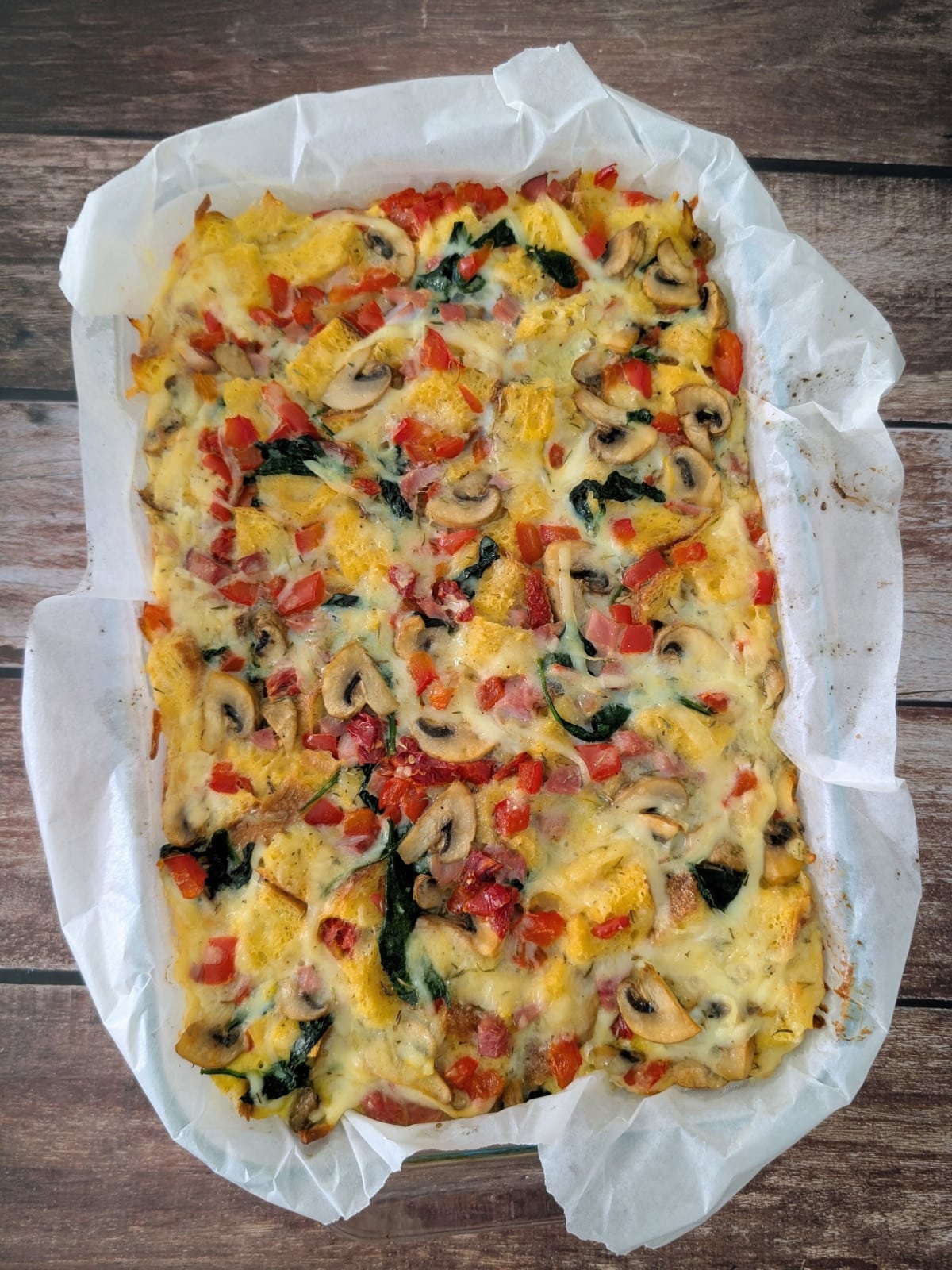 Golden breakfast strata with mushrooms, red peppers, spinach, ham and melted cheese in a parchment-lined baking dish.